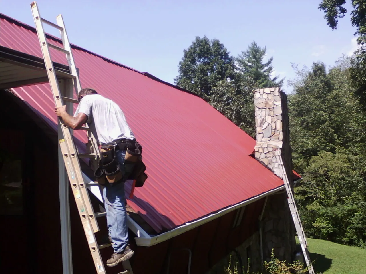 Roofer on a ladder beside a red metal roof and stone chimney — Soffit & Fascia Repair by Roofing Brothers in Trezevant